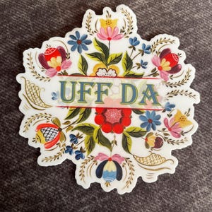 diecut adhesive sticker features colorful Norwegian rosemaling with the phrase ‘Uff Da’ written across the center