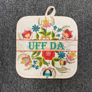 May include: Square, off-white pot holder with a floral design in red, blue, yellow, and pink. The text "UFF DA" is in teal. The pot holder has a loop for hanging and a beige border.