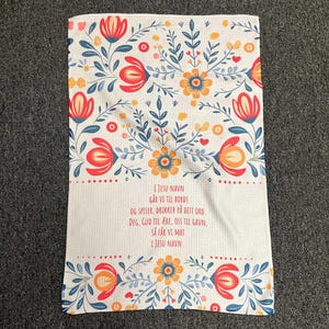 Puede incluir: Paño de cocina blanco con un diseño floral en rojo, naranja y azul. La toalla presenta un bloque de texto central con un verso religioso. El patrón floral incluye flores y hojas estilizadas.