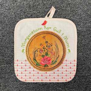 May include: Square beige pot holder with a loop for hanging. The center features a circular design with a floral pattern in pink, green, and yellow. The text "Morgenstunn har Gull i Munn" is printed around the circle.