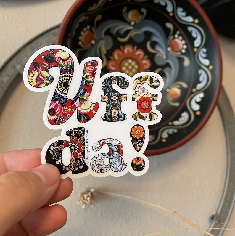 A white diecut sticker with the words "Uff da!" in a colorful, patterned font. Different colorful Norwegian rosemaling patterns create each letter.