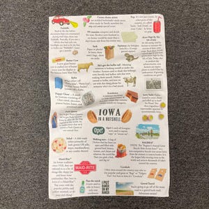 May include: White cotton kitchen towel with a colorful print featuring Iowa-themed illustrations and text. The towel includes images of food, landmarks, and phrases like "Iowa in a Nutshell" and "Maid-Rite."