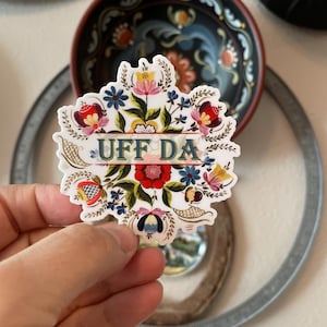 diecut adhesive sticker features colorful Norwegian rosemaling with the phrase ‘Uff Da’ written across the center