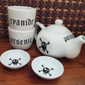 May include: A white ceramic teapot, two small cups, and two small bowls. The teapot has a skull and crossbones and the word "poison". The cups are labeled "cyanide" and "arsenic". The bowls have a skull and crossbones design. The set is on a wooden surface.