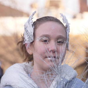 Pu&ograve; includere: Una giovane persona indossa una fascia per capelli con piume bianche e strass e un cappotto bianco con bordatura in pelliccia. La persona guarda la fotocamera e ha un trucco argentato glitterato sul viso. Lo sfondo &egrave; sfocato e sembra essere una strada cittadina.