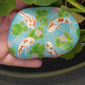 May include: A hand-painted, oval-shaped stone featuring a serene pond scene. The stone is painted with a light blue background, koi fish, lily pads, and small pink flowers. The koi fish have orange and white markings.