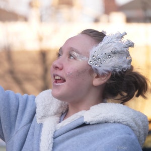 Pu&ograve; includere: Una giovane persona indossa una giacca blu chiaro con un colletto di pelliccia bianca e una fascia per capelli di piume bianche con strass argentati. La persona sta guardando in alto e sorridendo.
