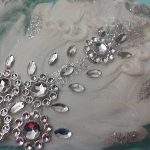 Pu&ograve; includere: Un cerchietto di piume bianche con glitter argentati e strass. Il cerchietto &egrave; decorato con un gruppo di grandi strass al centro e strass pi&ugrave; piccoli ai bordi.