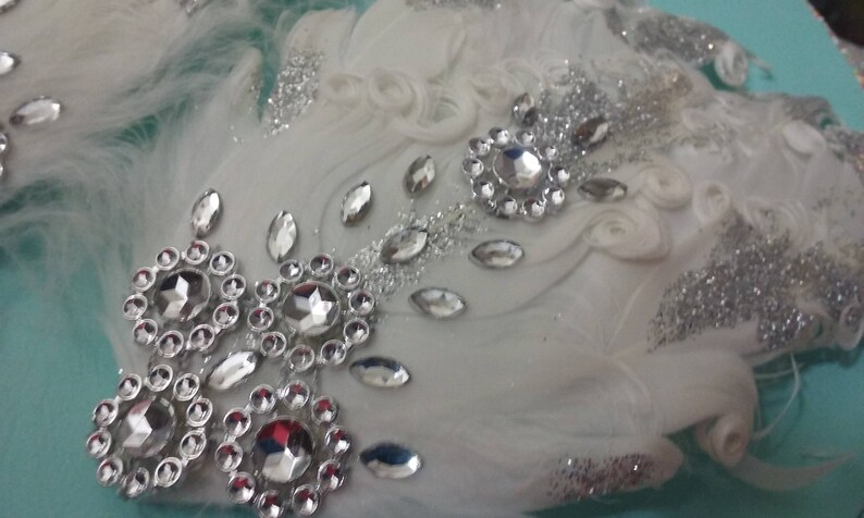 Pu&ograve; includere: Un cerchietto di piume bianche con glitter argentati e strass. Il cerchietto &egrave; decorato con un gruppo di grandi strass al centro e strass pi&ugrave; piccoli ai bordi.