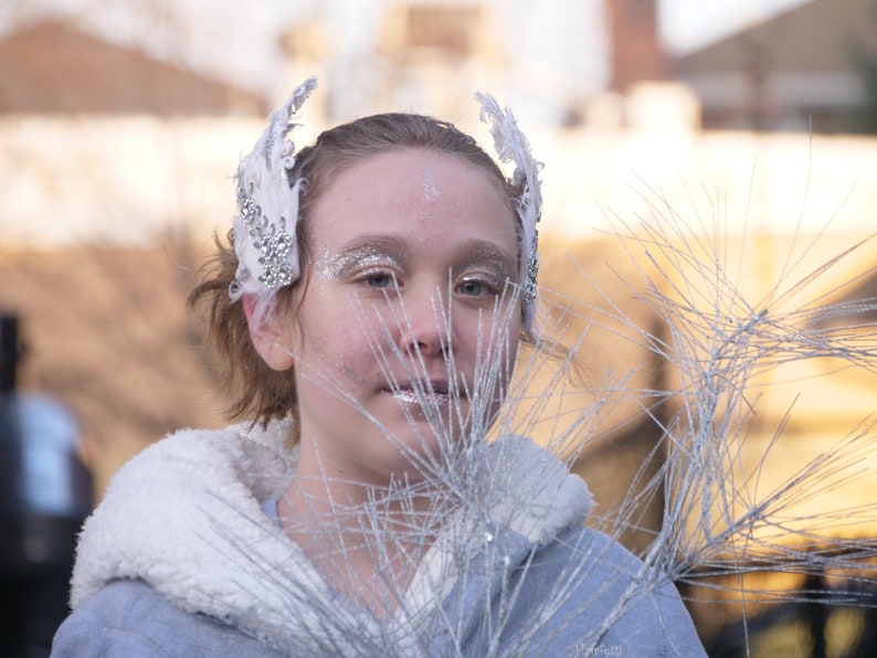 Pu&ograve; includere: Una giovane persona indossa una fascia per capelli con piume bianche e strass e un cappotto bianco con bordatura in pelliccia. La persona guarda la fotocamera e ha un trucco argentato glitterato sul viso. Lo sfondo &egrave; sfocato e sembra essere una strada cittadina.