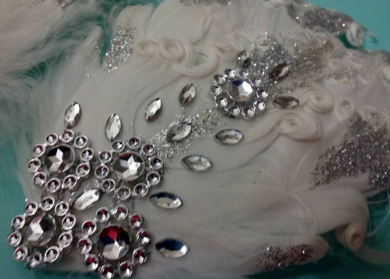Pu&ograve; includere: Fascia per capelli in piume bianche con glitter argentati e strass. La fascia &egrave; decorata con un grande gruppo di strass al centro e strass pi&ugrave; piccoli sparsi su tutta la superficie.