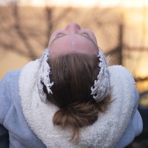 Pu&ograve; includere: Una donna con i capelli castani indossa una giacca blu con un colletto di pelliccia bianca. Sta guardando in alto a bocca aperta e ha un cerchietto di piume argentate e bianche nei capelli.