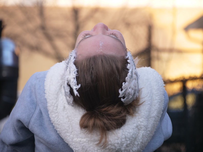 Pu&ograve; includere: Una donna con i capelli castani indossa una giacca blu con un colletto di pelliccia bianca. Sta guardando in alto a bocca aperta e ha un cerchietto di piume argentate e bianche nei capelli.