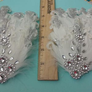 Pu&ograve; includere: Due fermagli per capelli in piume bianche con glitter argentati e strass trasparenti. I fermagli sono lunghi circa 10 cm.