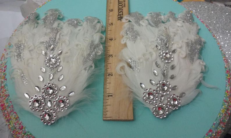 Pu&ograve; includere: Due fermagli per capelli in piume bianche con glitter argentati e strass trasparenti. I fermagli sono lunghi circa 10 cm.