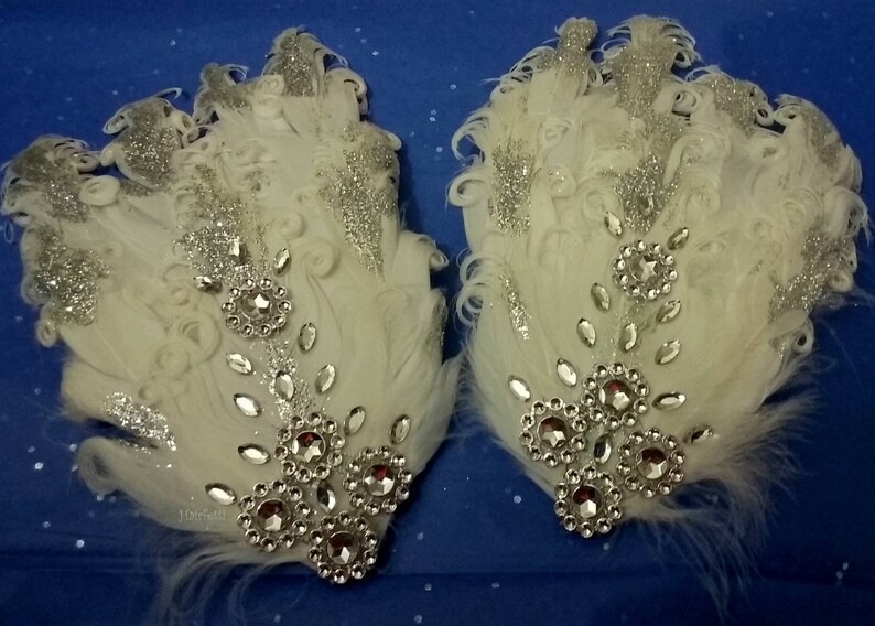 Pu&ograve; includere: Due fermagli per capelli in piume bianche con glitter argentati e strass. I fermagli sono decorati con un gruppo di strass al centro.