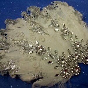 Pu&ograve; includere: Pettine per capelli in piume bianche con glitter argentato e strass. Il pettine &egrave; decorato con un gruppo di grandi strass al centro e strass pi&ugrave; piccoli sui bordi.