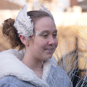 Pu&ograve; includere: Una giovane donna con un cerchietto di piume bianche adornato con strass. Indossa una giacca bianca con bordatura in pelliccia e ha un trucco glitterato argento sul viso.