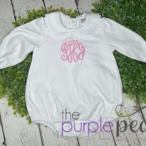 May include: White long-sleeved baby onesie with a ruffled collar and pink embroidered monogram. The onesie has a gathered bottom and is made of soft cotton.