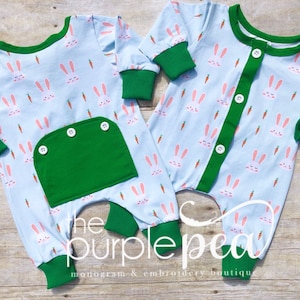 May include: Two baby onesies with a light blue background and a pattern of white bunnies and orange carrots. One onesie has a green pocket with three buttons. The other onesie has a green button placket. Both onesies have green trim around the neck, cuffs, and legs. The text "the purple pea monogram & embroidery boutique" is visible at the bottom of the image.