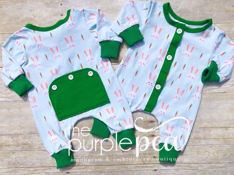 May include: Two baby onesies with a light blue background and a pattern of white bunnies and orange carrots. One onesie has a green pocket with three buttons. The other onesie has a green button placket. Both onesies have green trim around the neck, cuffs, and legs. The text "the purple pea monogram & embroidery boutique" is visible at the bottom of the image.