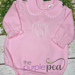 May include: Pink baby romper with a white scalloped collar and cuffs. Features a white embroidered monogram and the text "the purple pea monogram & embroidery boutique" in purple.