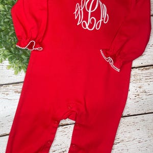 May include: Red baby onesie with white scalloped trim around the collar and cuffs. The onesie has a white embroidered monogram on the front.