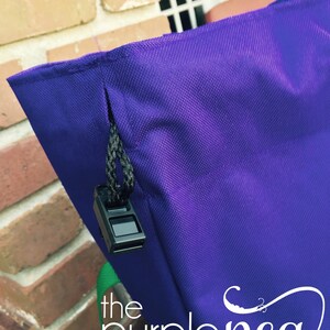 Puede incluir: Bolsa de mano morada con cierre de cremallera y un tirador de cremallera negro. La bolsa tiene una correa verde y el texto "the purple pea monogram & embroidery boutique" est&aacute; impreso en la parte inferior.