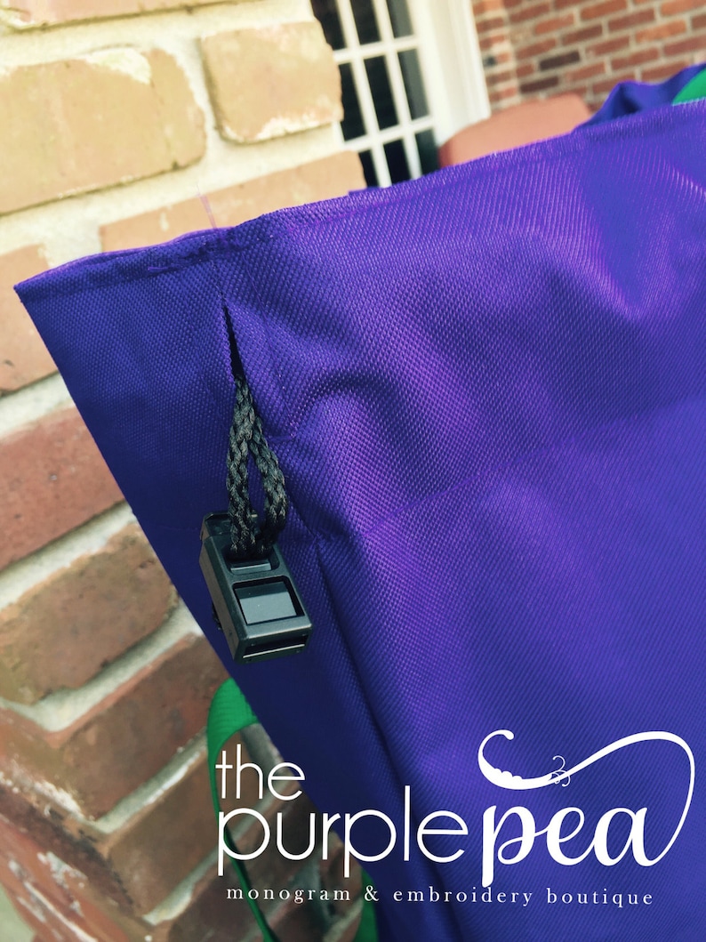 Puede incluir: Bolsa de mano morada con cierre de cremallera y un tirador de cremallera negro. La bolsa tiene una correa verde y el texto "the purple pea monogram & embroidery boutique" est&aacute; impreso en la parte inferior.