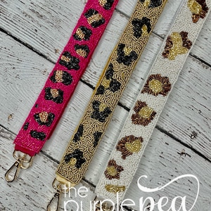 May include: Three beaded handbag straps with leopard print designs. The straps are pink, white, and brown with gold beads. The straps are shown on a white wood background. The text "the purple pea monogram & embroidery boutique" is at the bottom of the image.