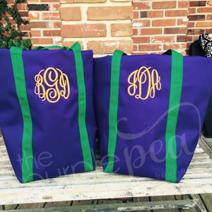 Puede incluir: Dos bolsas de mano de color morado con correas verdes y monogramas bordados en oro. Las bolsas est&aacute;n sobre una superficie de madera.