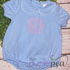 May include: Light blue baby romper with a Peter Pan collar trimmed with white scalloped edging. Features a pink monogram and the text "the purple pea" in purple. The romper has short sleeves and a gathered bottom.