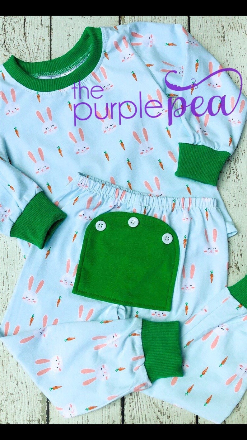 May include: A light blue pajama set with a bunny and carrot print. The set has green trim and a green pocket on the pants. The text "the purple pea" is visible on the top.