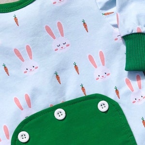May include: A light blue baby onesie with a green pocket and white buttons. The onesie has a repeating pattern of white bunnies and orange carrots. The text "the purple pea" is printed on the pocket.