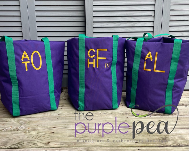 Puede incluir: Tres bolsas de lona moradas con correas verdes y iniciales bordadas en amarillo. Las bolsas est&aacute;n sobre una superficie de madera con el texto "the purple pea monogram & embroidery boutique" debajo.