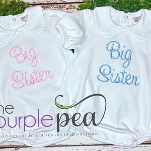 May include: Two white baby rompers with Peter Pan collars. One has pink embroidered text reading "Big Sister," the other has blue text. The logo "the purple pea" is at the bottom. Monogram & embroidery boutique.
