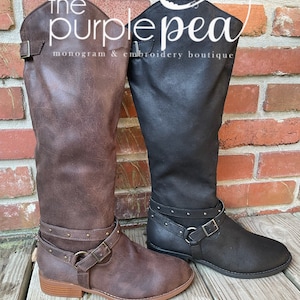 May include: Two pairs of brown and black knee-high boots with buckles and studs. The boots are on a wooden surface with a brick wall in the background. The text "the purple pea monogram & embroidery boutique" is visible on the wall.