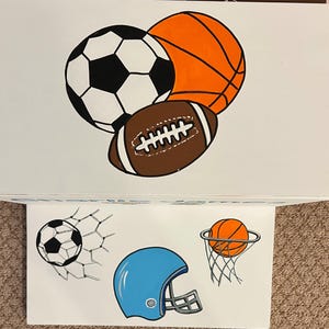 May include: A white wooden box with a painted design of a soccer ball, basketball, and football. The box is open and shows a white surface with a painted design of a soccer ball in a net, a basketball in a hoop, and a blue football helmet.