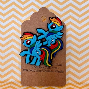May include: Two blue and rainbow-colored pony charms with wings and a rainbow tail. The charms are on a brown tag with the text "The Geek Collective" and "etsy.com/shop/ChaseAndCharlie".