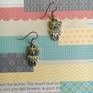 May include: A pair of silver owl earrings with a detailed design. The owls have large eyes and are hanging from silver hooks.