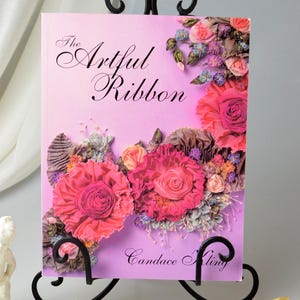 May include: A book titled 'The Artful Ribbon' by Candace Kling. The book cover features a pink background with a floral design made of ribbon. The book is displayed on a black metal book stand.