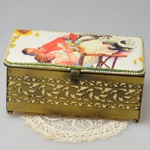 May include: A vintage brass jewelry box with a hinged lid, featuring a decorative floral design on the sides and a painted scene on the top. A white flower, gold ribbon, and pearl necklace are next to the box.