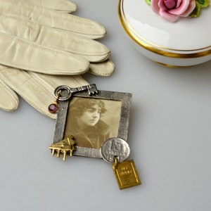May include: A silver-toned picture frame with a black and white photo of a young woman. The frame is decorated with a gold key, a small pink gem, a gold piano, and a gold coin with the number 15 on it. The frame is hanging from a gold chain with a small gold book charm.