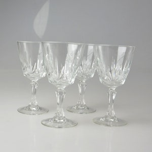 May include: Four clear crystal wine glasses with intricate starburst patterns on the bowls. The glasses have slender stems and round bases. The glasses are arranged in a row on a white surface.