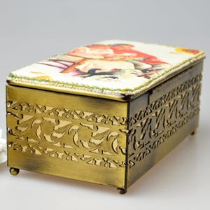 May include: Rectangular antique brass jewelry box with a decorative floral design on the sides. The lid features a colorful illustration. The box is set on small feet and has a beaded trim.