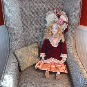 May include: A porcelain doll wearing a pink and white dress with a red velvet jacket sits in a gray armchair with a patterned fabric. The doll has blonde hair and blue eyes and is wearing a white hat with a pink flower.