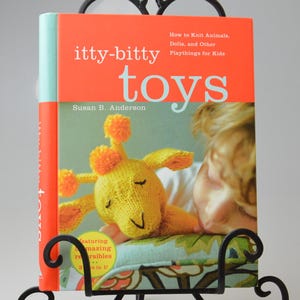 Op de afbeelding: Een zwarte metalen boekenstandaard met een boek getiteld "itty-bitty toys" met een gebreide giraf op de cover. Een kleine, gehaakte aardbei- en bladdecoratie staat ervoor.