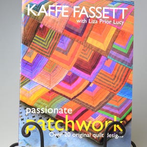 May include: A book titled "KAFFE FASSETT" with "passionate patchwork" on the cover, displayed on a black metal stand. The book cover features a colourful patchwork design in shades of orange, purple, and green. The stand has decorative scrollwork.