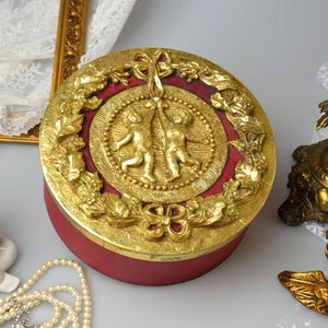 May include: A round, red and gold trinket box with a raised design of two cherubs holding a ribbon. The box is decorated with gold leaf and has a floral motif.