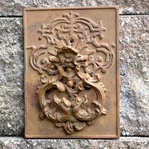 May include: A rusty metal door knocker with an ornate design. The knocker is shaped like a pair of stylized birds with their heads facing each other. The knocker is mounted on a rectangular metal plate.